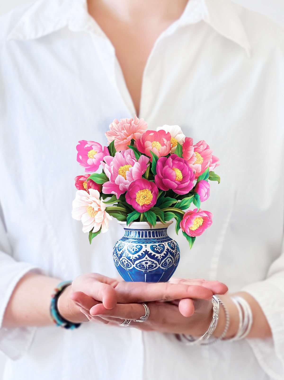 A person in a white shirt is holding the FreshCut Paper's Mini Peony Paradise, showcasing vibrant pink and white flowers. The person's head is not visible, and they are adorned with bracelets on both wrists. The attention is captivated by the meticulously detailed and colorful flower arrangement that exudes classic sentiments.
